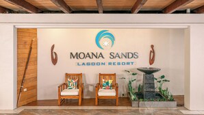 Uma área de recepção com teto de madeira, duas cadeiras de madeira com almofadas coloridas, um logotipo acima do nome 'MOANA SANDS LAGOON RESORT' e uma pequena fonte com plantas.