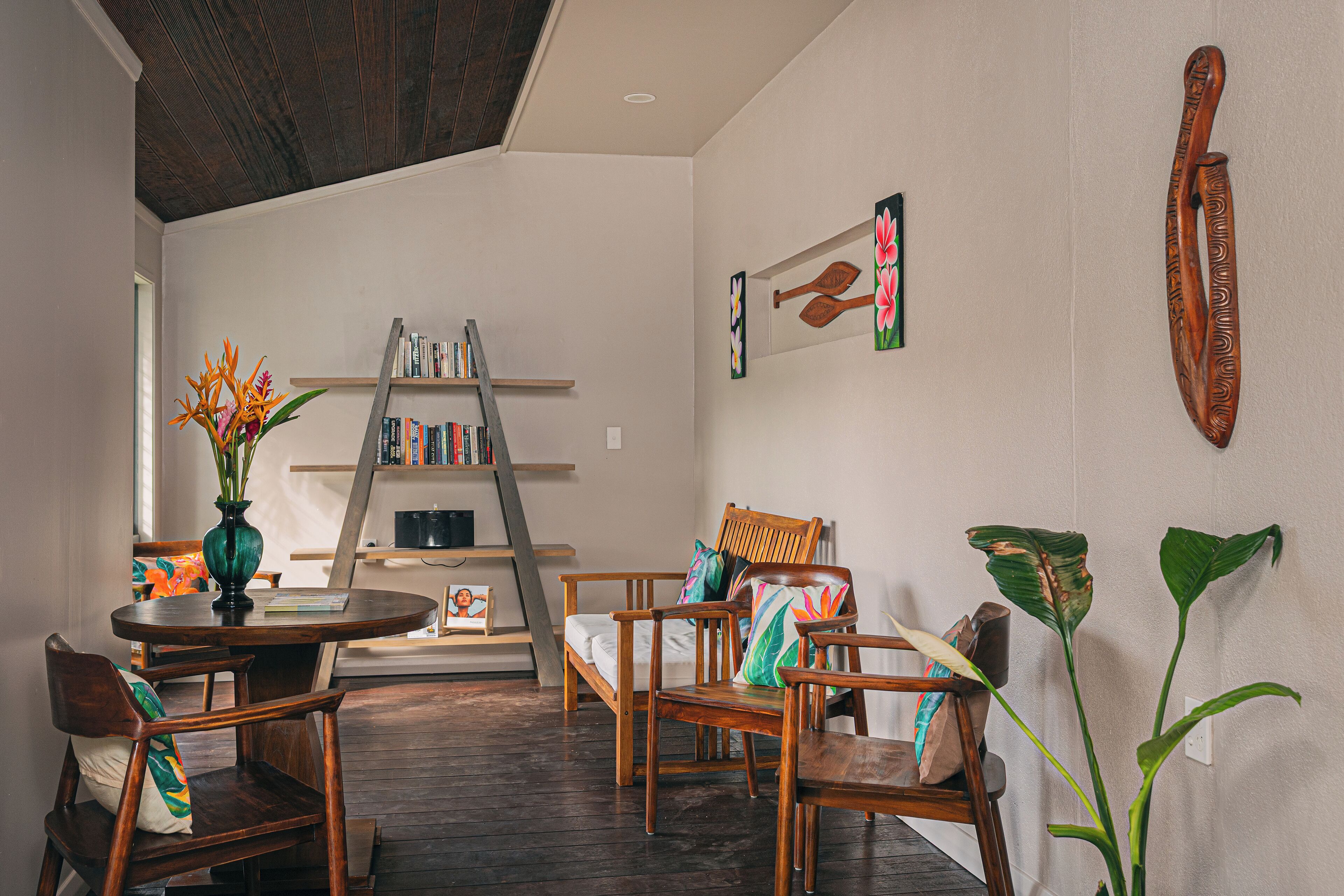 A cozy living room with a wooden coffee table, two wooden chairs, a bookshelf, and a large plant.