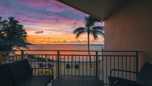 Uma varanda com vista para o pôr do sol sobre o oceano, uma palmeira e uma praia com algumas cadeiras e uma mesa.