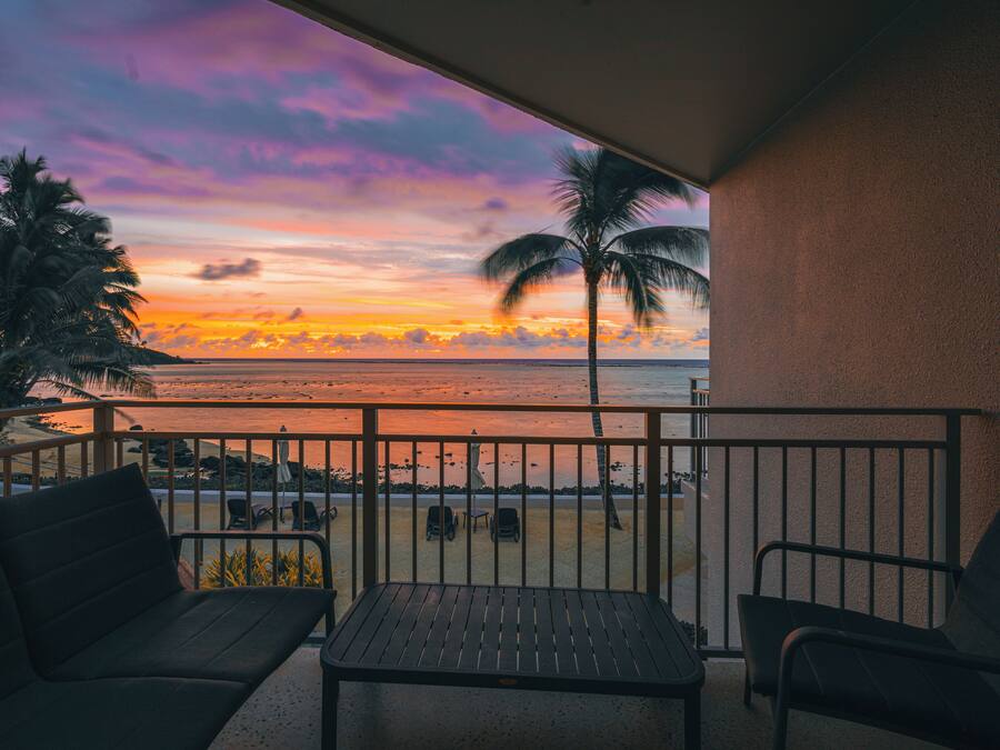 Un balcón con vista al atardecer sobre el océano, una palmera y una playa con algunas sillas y una mesa.