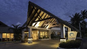Entrada de um resort com telhado de palha, uma placa com a inscrição 'Moana Sands Resort' e um caminho bem iluminado que leva ao edifício.