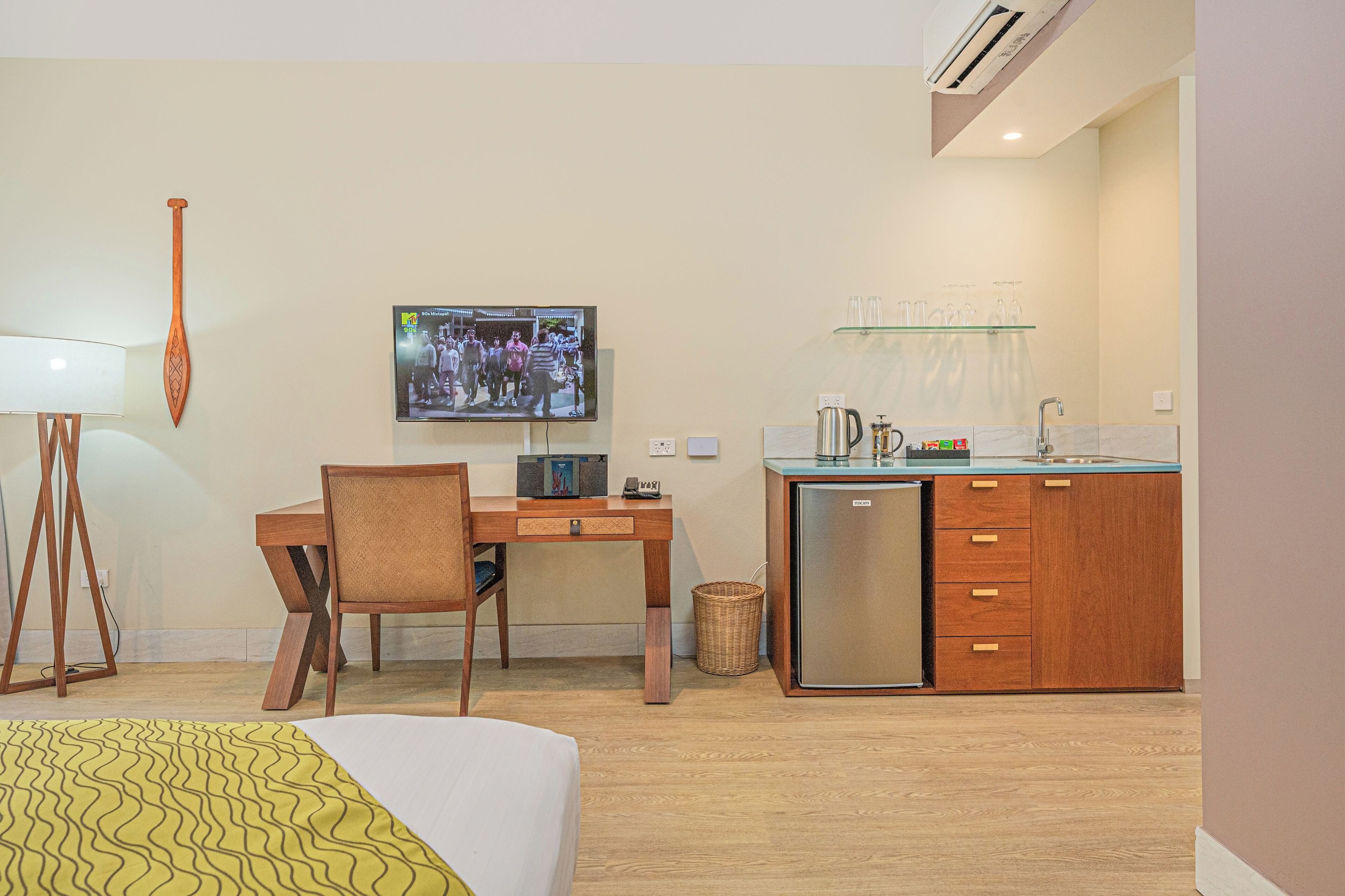 Ein Hotelzimmer mit einem Bett, einem Schreibtisch mit Stuhl, einem an der Wand befestigten Fernseher, einer Küchenzeile mit Kühlschrank und Spüle sowie einem kleinen Mülleimer.