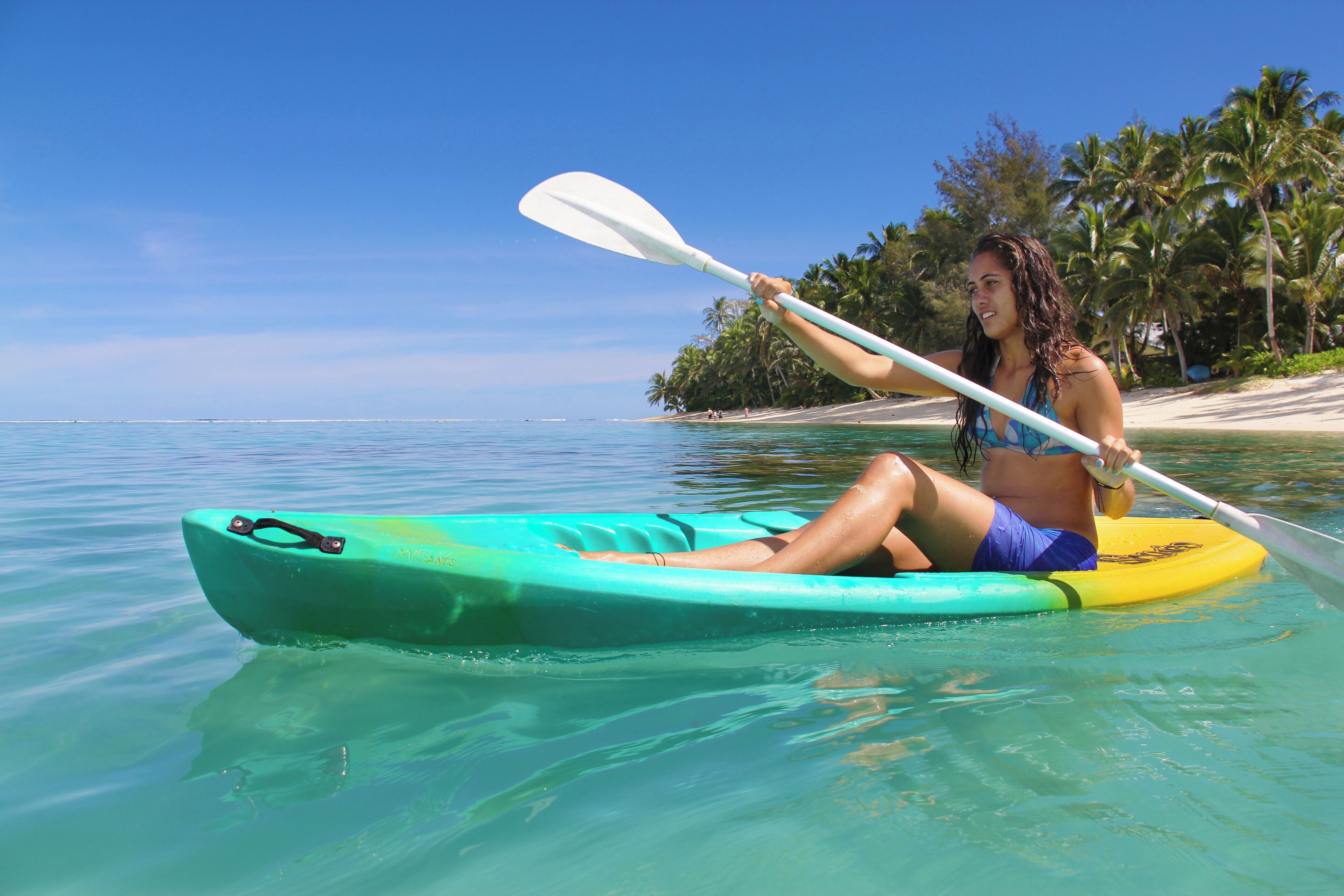 A person kayaking on a clear day with a beach and palm trees in the background.
