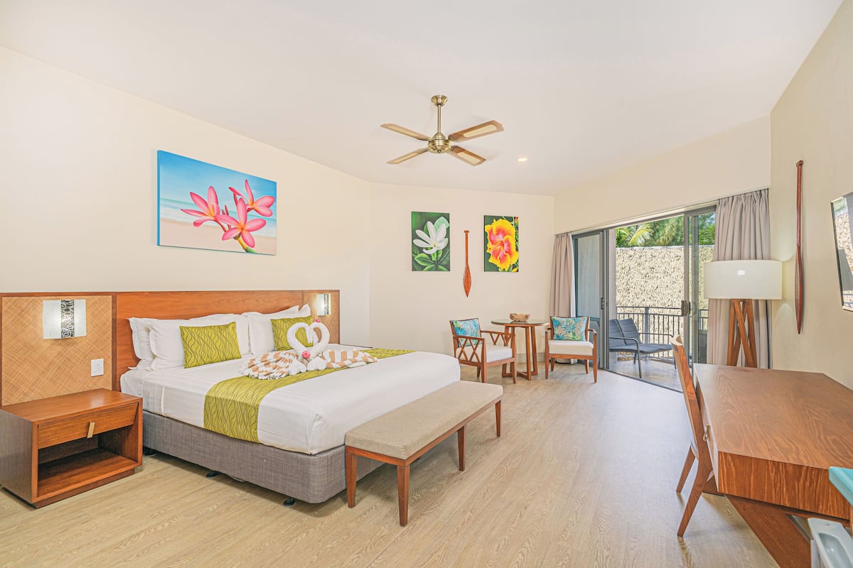 A hotel room with a bed, a desk, a chair, a bench, a ceiling fan, and artwork on the walls.