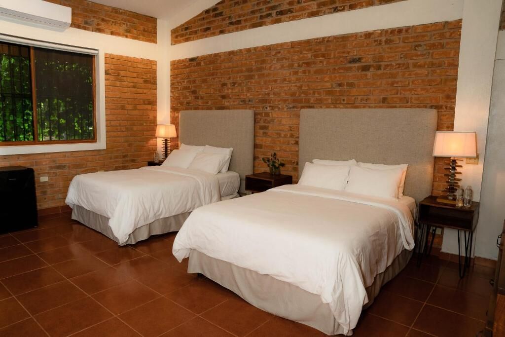 Habitación de hotel con dos camas, una pared de ladrillo, un ventanal con vistas y una mesita con lámpara.