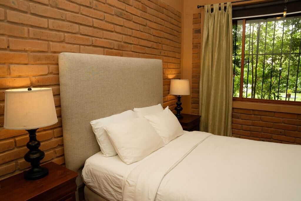 A bed with white bedding, two bedside lamps, a brick wall, and a window with curtains.