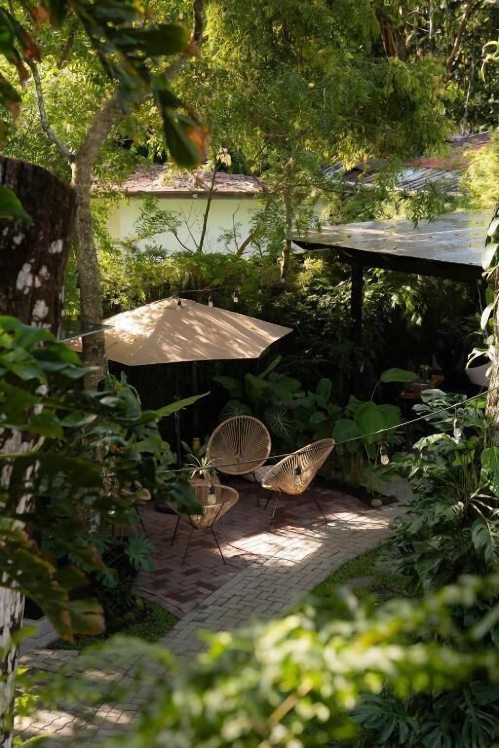 Un patio exterior con dos sillas de mimbre y un paraguas grande, rodeado de abundante vegetación y un camino de ladrillo.