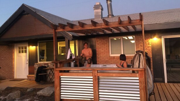 Een houten terras met een pergola, een huis met bakstenen muren en een schoorsteen, en een schuifdeur met ruit.'