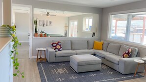 Een moderne woonkamer met een grijze modulaire bank, een klein voetenbankje en een kamerkleed met een patroon. Aan de muur hangt een grote spiegel, er staat een pot met een plant en er is een raam met jaloezieën.