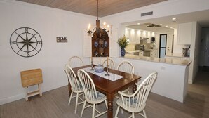 A dining area with a wooden table, white chairs, a compass wall decoration, and a kitchen in the background. - The Shore 211-Stylish 2BR Gulffront Condo with Stunning Sunsets & Pool by RVA (Longboat Key)