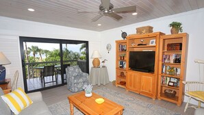 A living room with a TV, wooden bookshelves, a ceiling fan, and a view through a glass door. - The Shore 211-Stylish 2BR Gulffront Condo with Stunning Sunsets & Pool by RVA (Longboat Key)