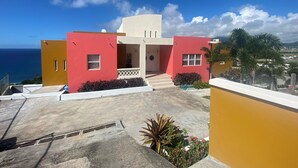 Un bâtiment moderne à la façade rouge et jaune, doté d’un balcon orné de plantes et offrant une vue sur l’océan.