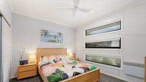 Ein modernes Schlafzimmer mit einem hölzernen Bett, einem Nachttisch mit Lampe, einem Deckenventilator und einem Strandbild an der Wand.