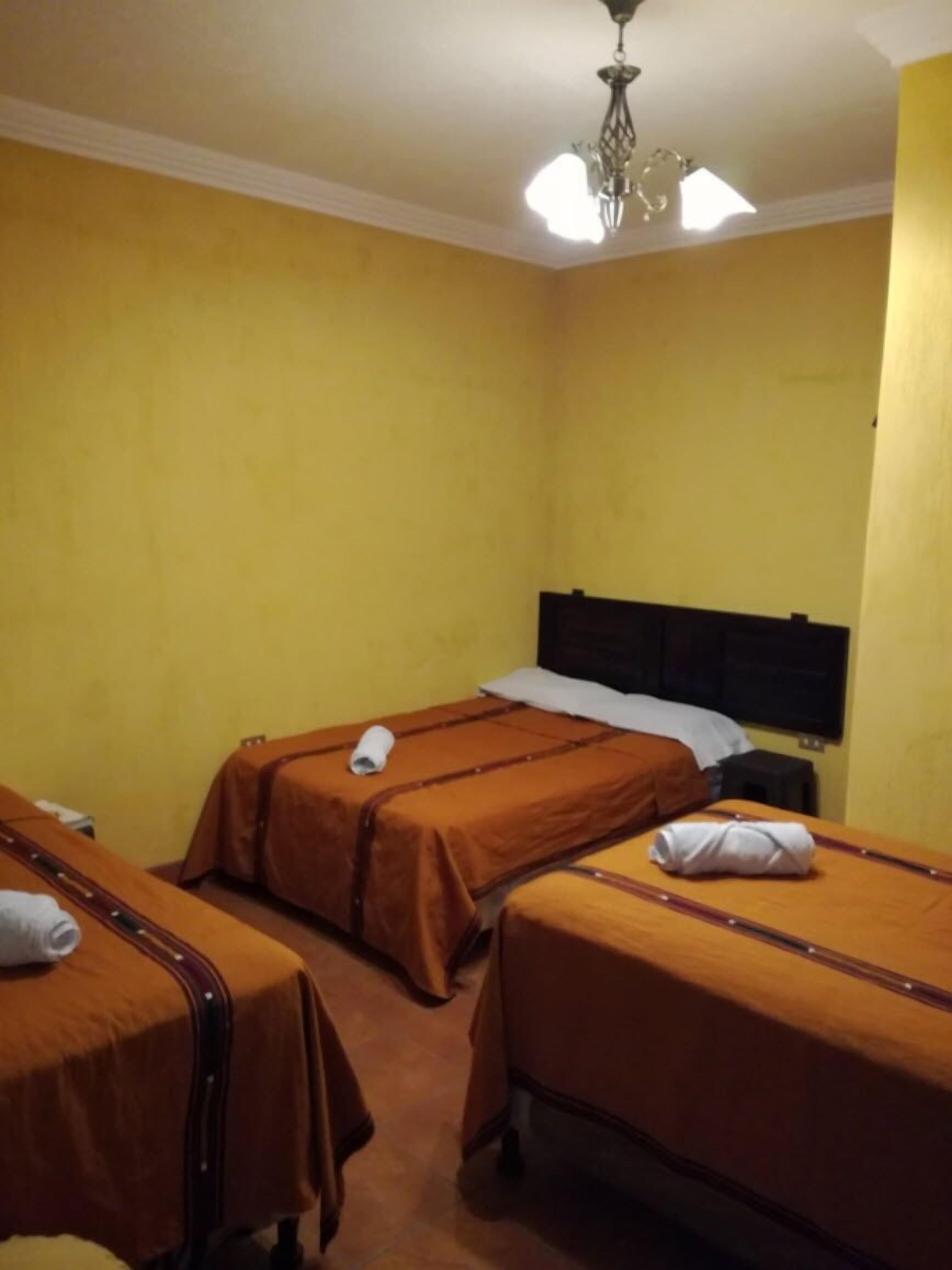 A hotel room with three single beds, each with a brown bedspread and white pillows. The room has yellow walls and a white ceiling with a chandelier.