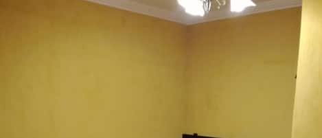 A hotel room with three single beds, each with a brown bedspread and white pillows. The room has yellow walls and a white ceiling with a chandelier.