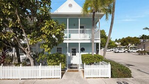 A two-story house with a balcony, surrounded by a white picket fence, palm trees, and a clear sky.