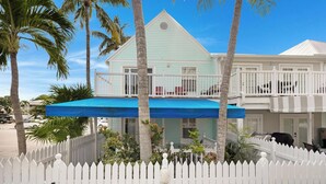 A two-story building with a white picket fence, a blue awning, and a balcony with red chairs.