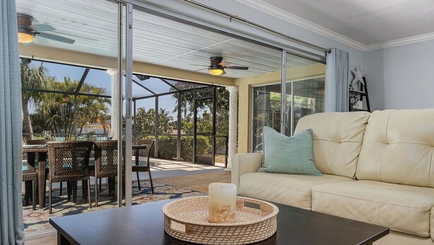 Ein Wohnzimmer mit einem beigefarbenen Sofa, einem Couchtisch mit einer Kerze, Korbstühlen und einer Glasdoor, die zu einem Außenbereich mit einem Pool führt.