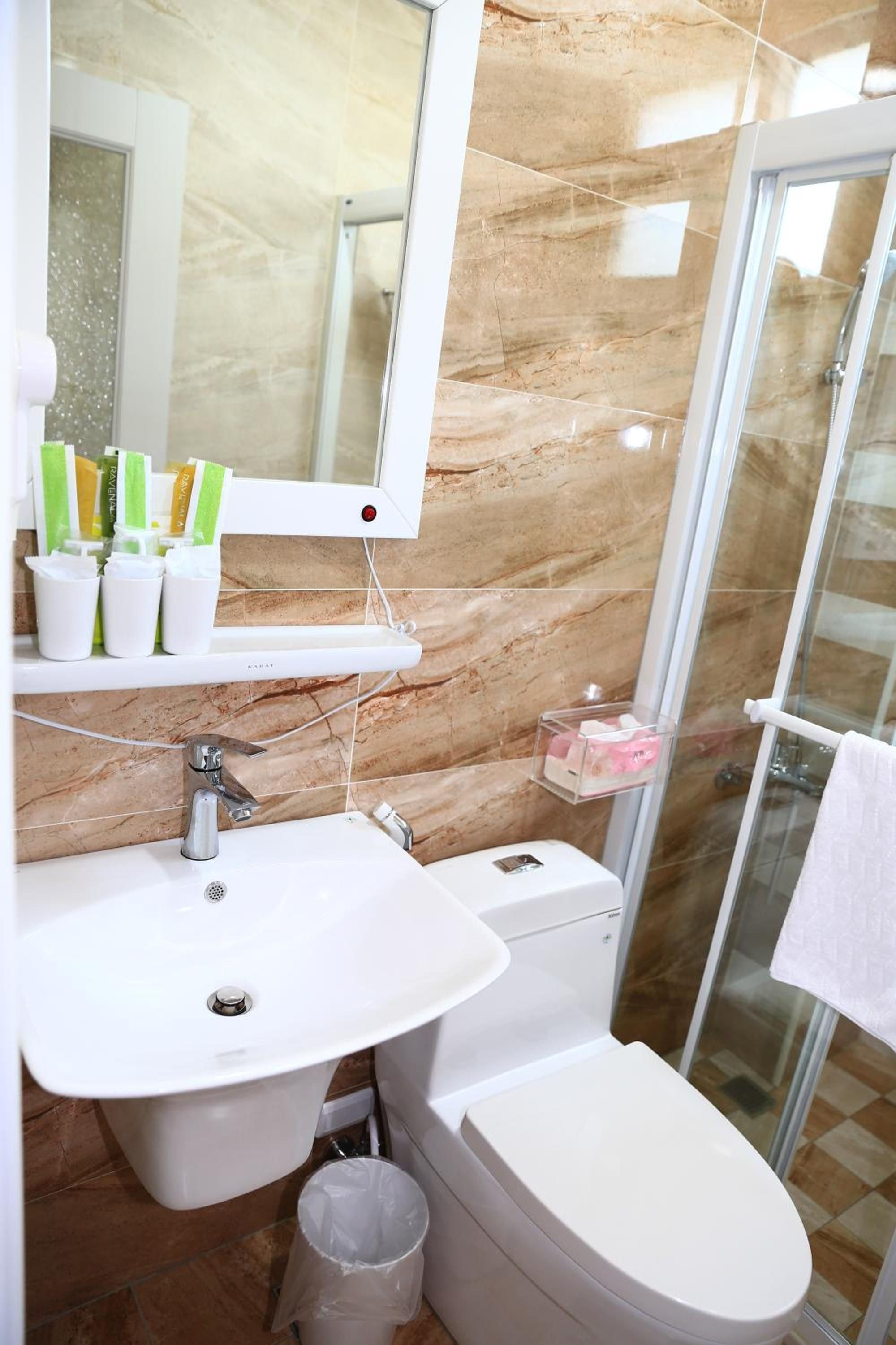 Un baño con lavamanos blanco, espejo y retrete. Hay artículos de aseo en el lavamanos y un cubo de basura.