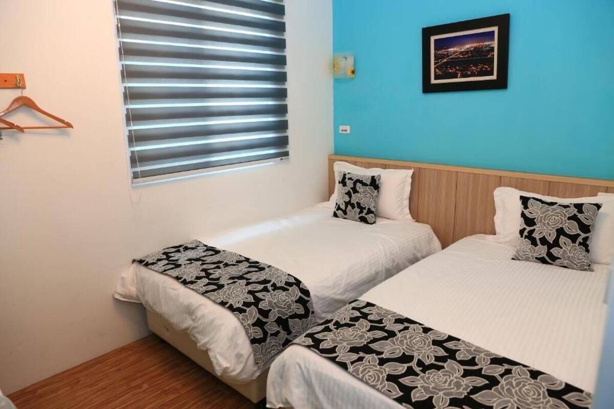A room with two beds, a window with blinds, a blue wall, and a framed picture.