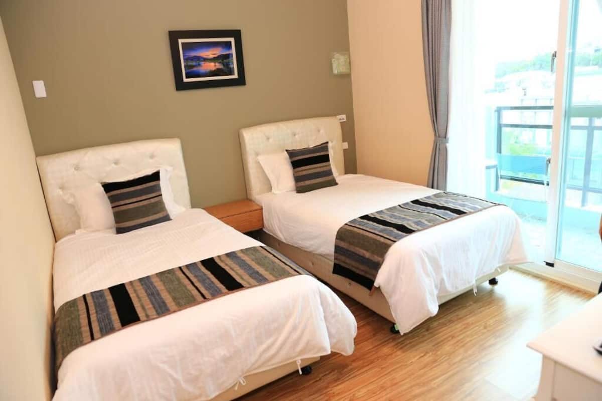 A hotel room with two beds, a small bedside table, a framed picture on the wall, and a balcony with a view.