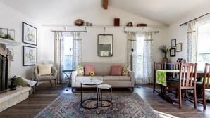 Una sala de estar con sofá, mesa de centro y zona de comedor con sillas. Cuenta con chimenea, espejo y alfombra.