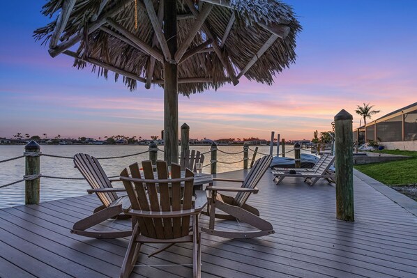 A wooden deck with Adirondack chairs, a thatched umbrella, and a view of the water at sunset. - Heated Pool, Beautiful Sunsets, Fishing Dock- Villa Thunderbird View - Roelens Vacations (Cape Coral)