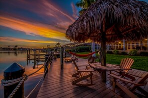 A wooden deck with rocking chairs and a hammock, overlooking a body of water and a sunset. - Heated Pool, Beautiful Sunsets, Fishing Dock- Villa Thunderbird View - Roelens Vacations (Cape Coral)