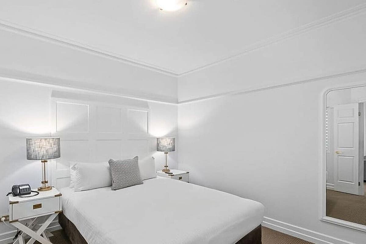 A white-themed bedroom with a large bed, two bedside tables with lamps, and a mirror on the wall.