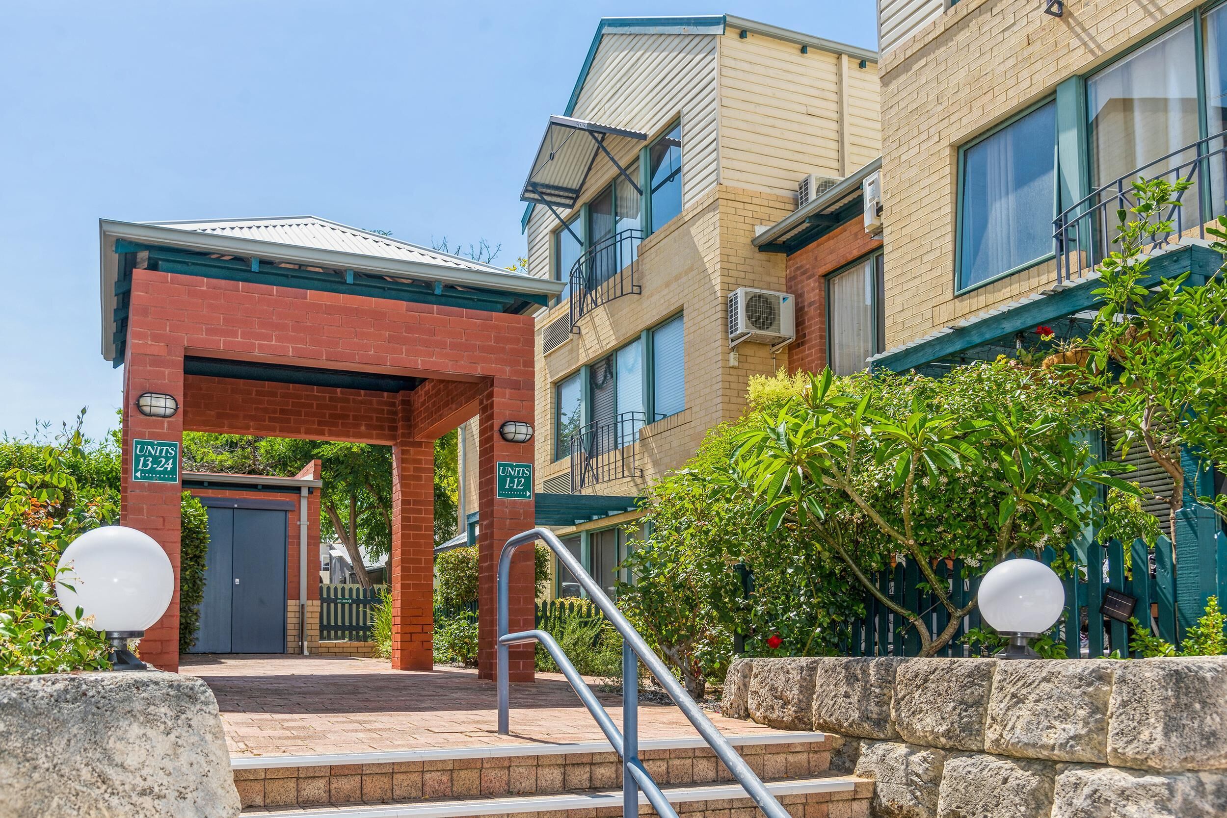Entrance to the apartment complex with brick archway, metal handrails, stone retaining walls, and lush green landscaping.