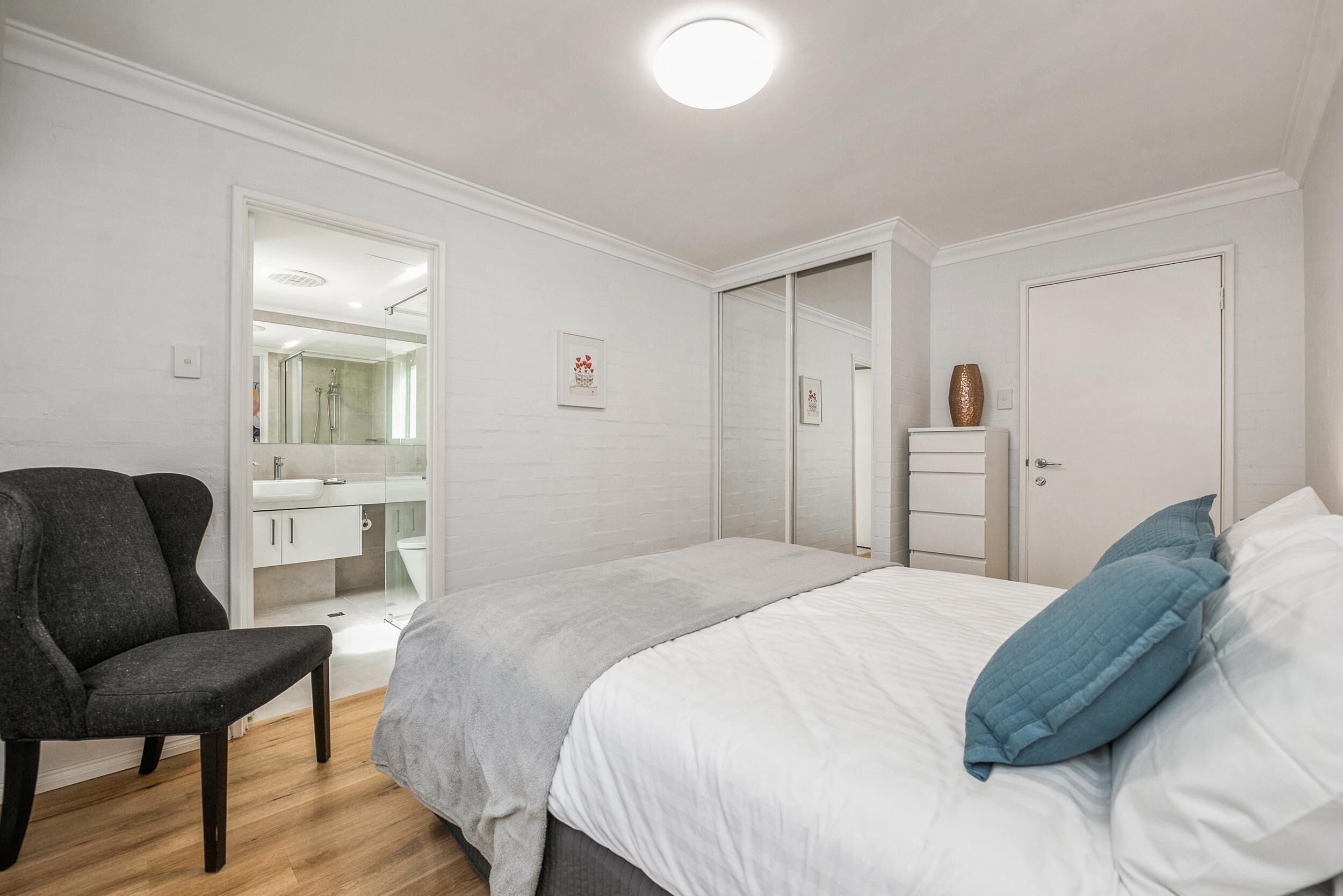 Bedroom with white linens, blue pillows, wood flooring, black armchair, mirrored closet doors, and ensuite bathroom.