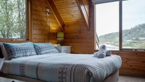 Un dormitorio con techo de madera, una cama con ropa de cama estampada, una mesita de noche con lámpara y vistas a un paisaje a través de un amplio ventanal.
