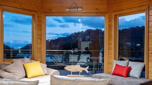 Interior acogedor de una cabaña con sofá y cojines, una decoración de pez de madera y ventanas grandes que ofrecen vistas del exterior.