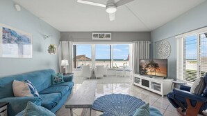 A living room with a blue sofa, a coffee table, a TV, and a view of the beach through large windows. - Island House Beach Resort 1S (Siesta key)