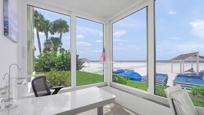 A room with a large window overlooking a beach, a white desk, and a wicker chair. - Island House Beach Resort 1S (Siesta key)