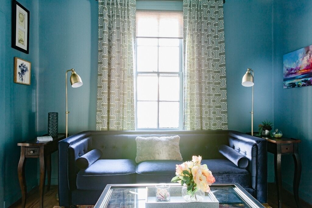 Una habitación con un sofá azul, una mesa de centro de vidrio con un jarrón, dos lámparas y cortinas estampadas.