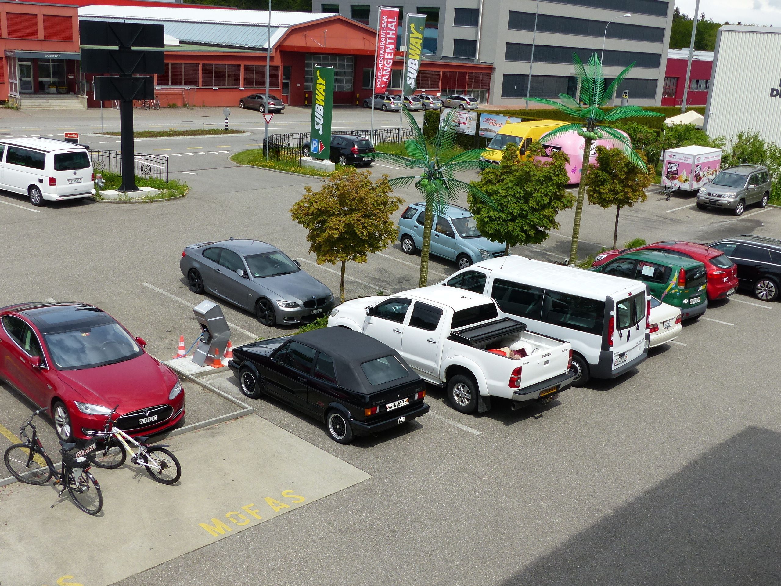 Un parking où sont garés divers véhicules, notamment des voitures, des fourgonnettes et une moto. On aperçoit des arbres et un bâtiment avec une enseigne Subway à l’arrière-plan.
