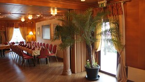 A dining area with a long table set for a meal, surrounded by chairs. There are potted plants and a large column in the center. - Hotel Langenthal (Langenthal)