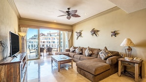 A spacious living room with a large sofa, a coffee table, and a TV. There are decorative wall pieces and a ceiling fan. - Beachfront 4th Floor with Stunning Views to Lands End, Medano Beach and Cabo San Lucas Bay (Cabo San Lucas)