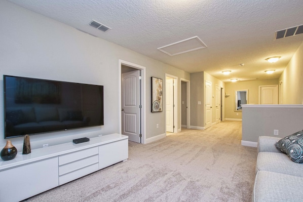 A modern living room with a flat-screen TV on a white entertainment unit, a grey sofa, and a hallway leading to other rooms.
