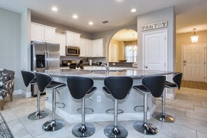 A modern kitchen with white cabinets, a stainless steel refrigerator, a built-in microwave, and a large island with black bar stools. - 6BR Solterra Pool Home | Spa, Game Room, BBQ, Themed Rooms & 3 Kings (Davenport)