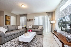 A living room with two grey sofas, a white coffee table, and a patterned rug. There is a flat-screen TV on the wall, a lamp, and a window with blinds. - 6BR Solterra Pool Home | Spa, Game Room, BBQ, Themed Rooms & 3 Kings (Davenport)