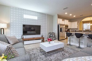A modern living room with a sofa, coffee table, and TV. There is a kitchen area with a refrigerator, microwave, and bar stools. - 6BR Solterra Pool Home | Spa, Game Room, BBQ, Themed Rooms & 3 Kings (Davenport)
