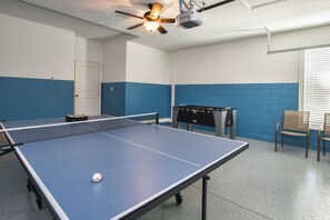 A ping pong table with a ball, a ceiling fan, and a foosball table in a room with blue and white walls.