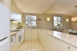 A kitchen with white cabinets, a stove, and a sink. There are windows with views of trees and a wall-mounted light fixture.