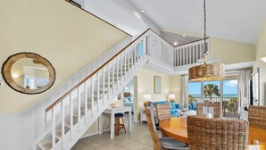 A spacious living area with a dining table surrounded by wicker chairs, a white staircase, and a large mirror on the wall. - Stay and Play all Day in this Very Nice Spacious Beach Resort Villa! C1622A (Englewood)