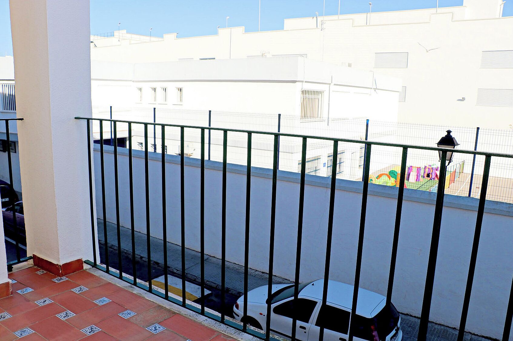 A balcony with a metal railing, a tiled floor, and a view of a street with buildings and a playground.