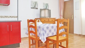 Une petite cuisine avec un meuble rouge, une table à manger en bois recouverte d’une nappe à carreaux et deux chaises. Deux tableaux encadrés ornent le mur, et une porte est dotée d’un rideau à rayures.