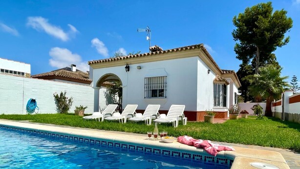 A poolside area with a clear blue pool, white lounge chairs, and a white building with a tiled roof.