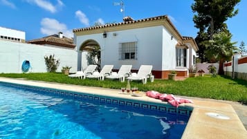 Un espace aménagé au bord d’une piscine à l’eau d’un bleu limpide, avec des chaises longues blanches et un bâtiment blanc doté d’un toit recouvert de tuiles.